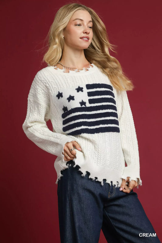 Woman wearing a cream distressed American flag sweater with raw hem and dark jeans against a red background