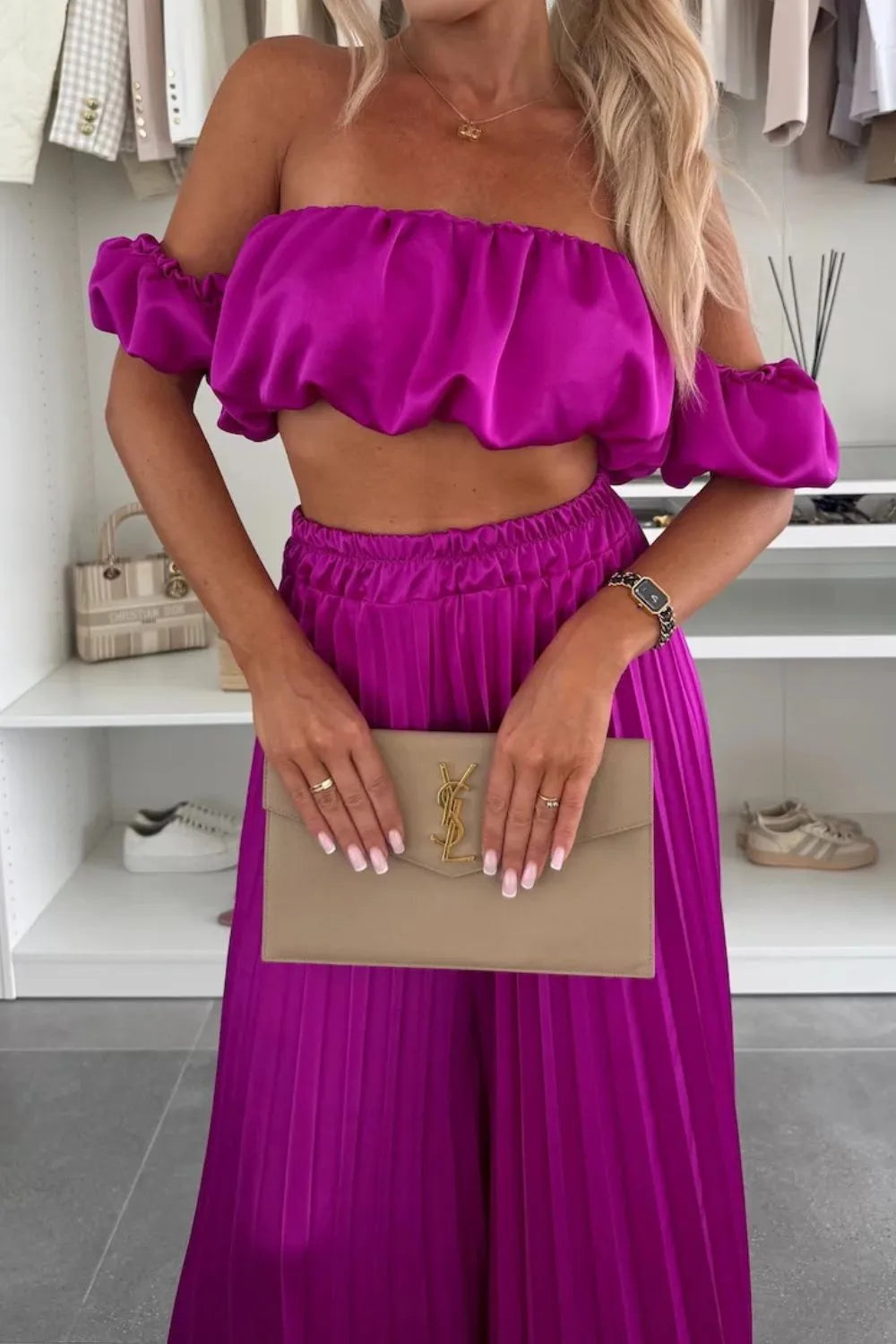 Woman wearing purple off-shoulder ruffled top and pleated pants set holding beige clutch