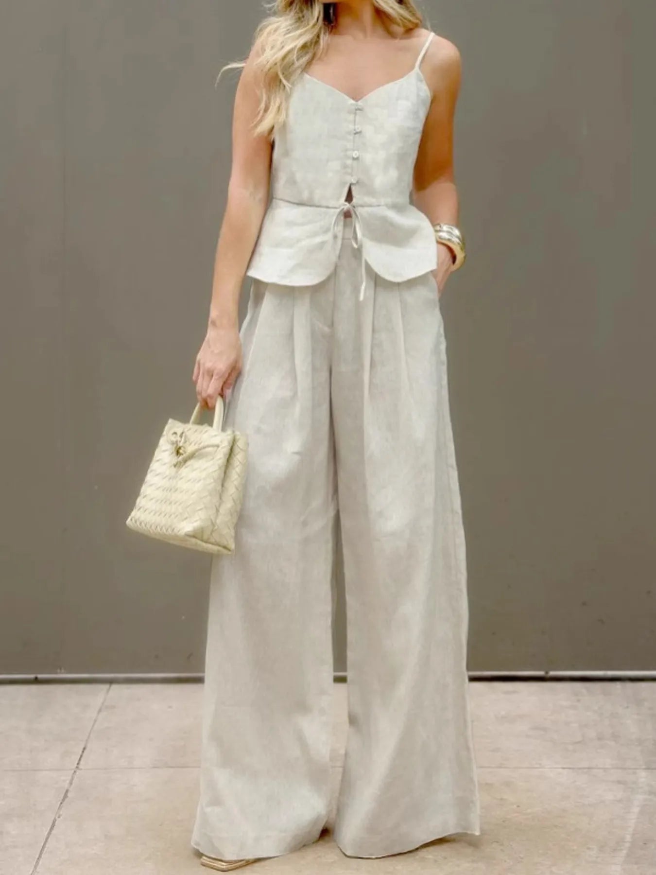 Woman wearing beige 100% cotton V-Neck Cami and Pants Set with buttoned tied top and wide-leg pants, holding woven handbag.