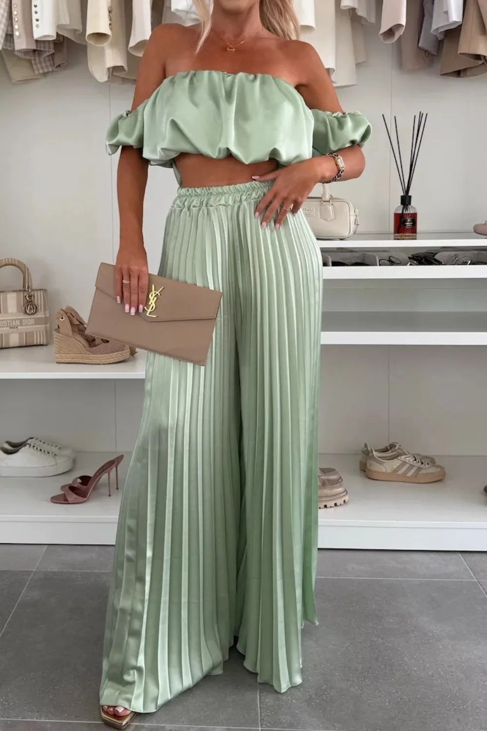 Woman wearing light green off-shoulder ruffled top and pleated pants set holding a beige clutch purse
