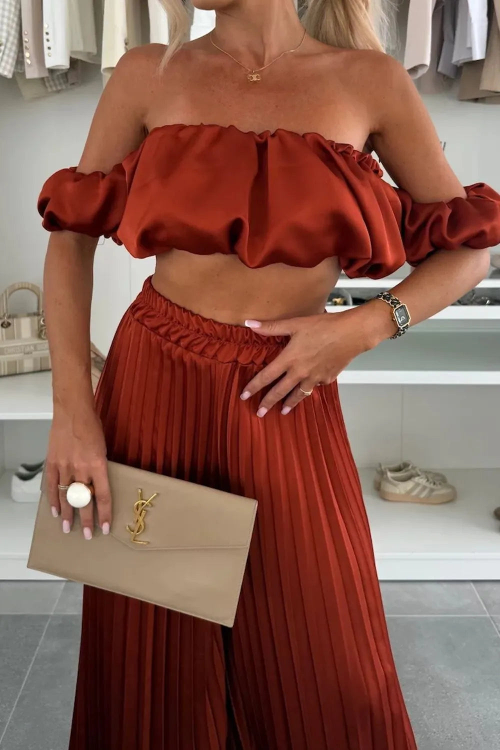 Rust-colored off-shoulder top with ruffled sleeves and pleated matching pants set holding beige clutch