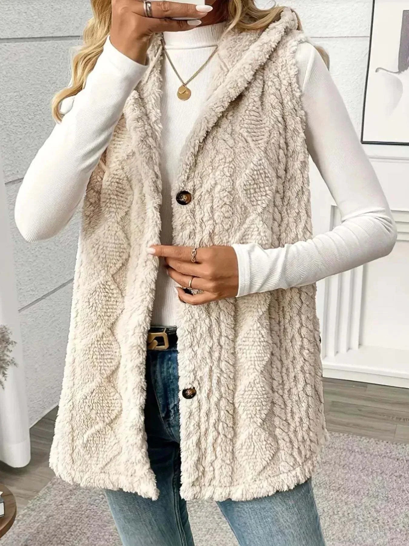 Beige button up plush hooded vest with textured knit pattern, sleeveless design, and front buttons worn over white long-sleeve top