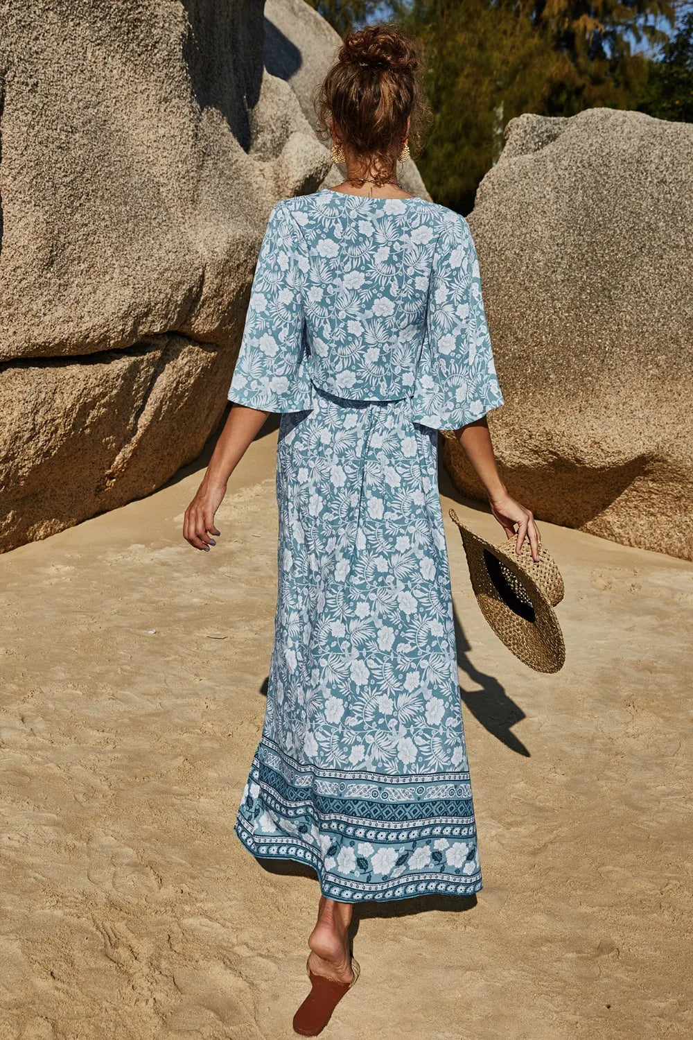 Woman wearing blue printed half sleeve top and slit skirt set walking on sandy beach holding straw hat