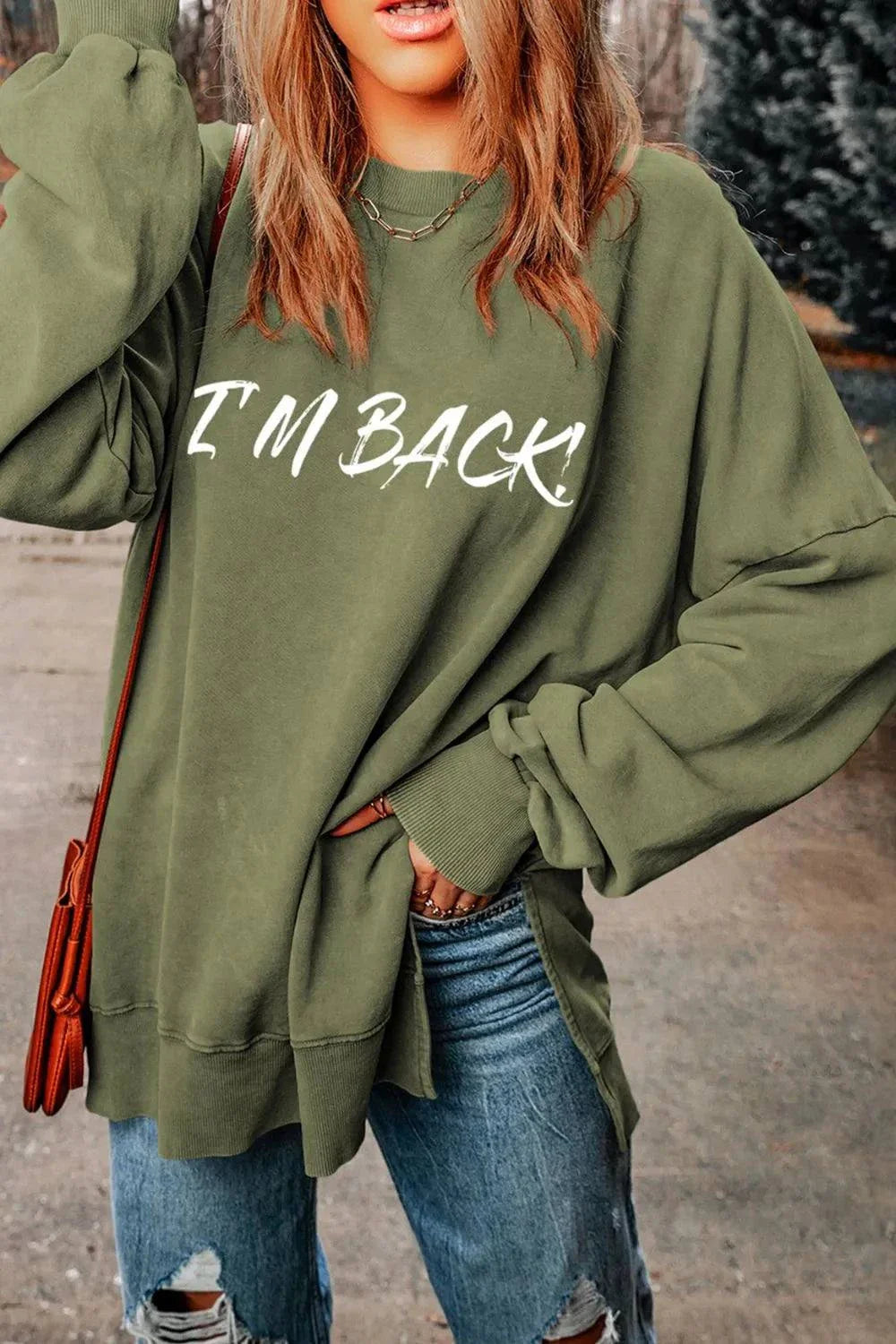Woman wearing green dropped shoulder sweatshirt with "I'M BACK" text and distressed jeans outdoors