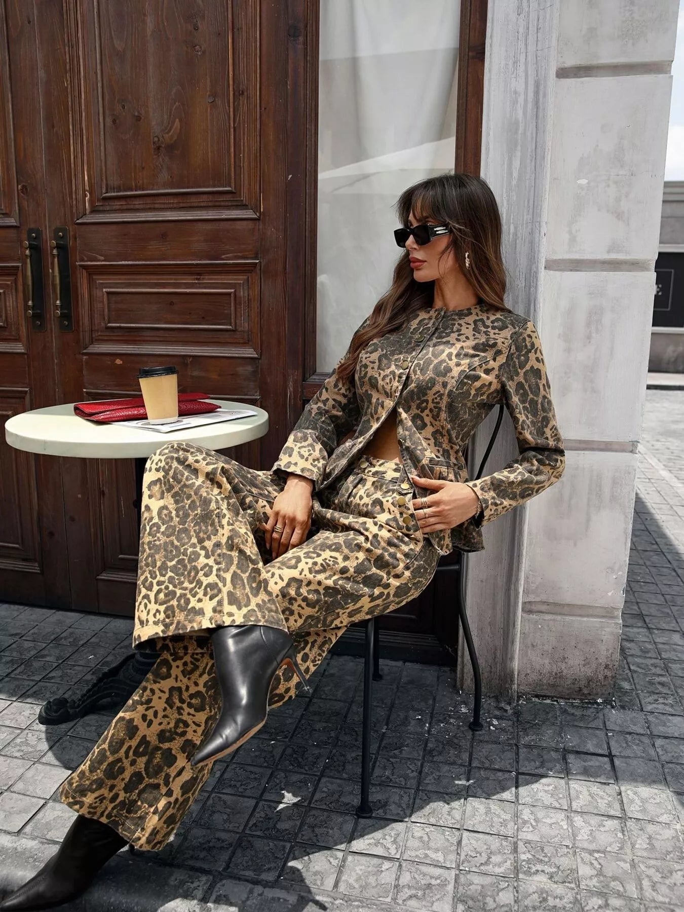Woman wearing leopard print button up long sleeve top and matching jeans denim set sitting outdoors with black boots and sunglasses
