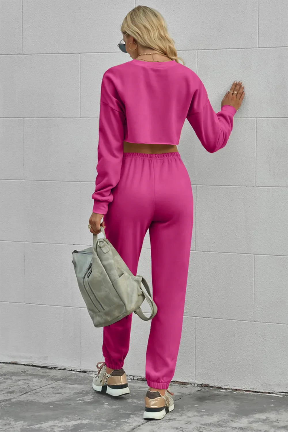 Woman wearing bright pink round neck long sleeve cropped top and pants set with drawstring waist, holding a gray backpack.