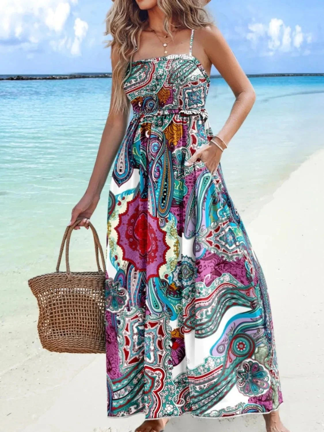 Woman wearing a colorful printed maxi cami dress with smocked waist detail on a beach, holding a woven handbag