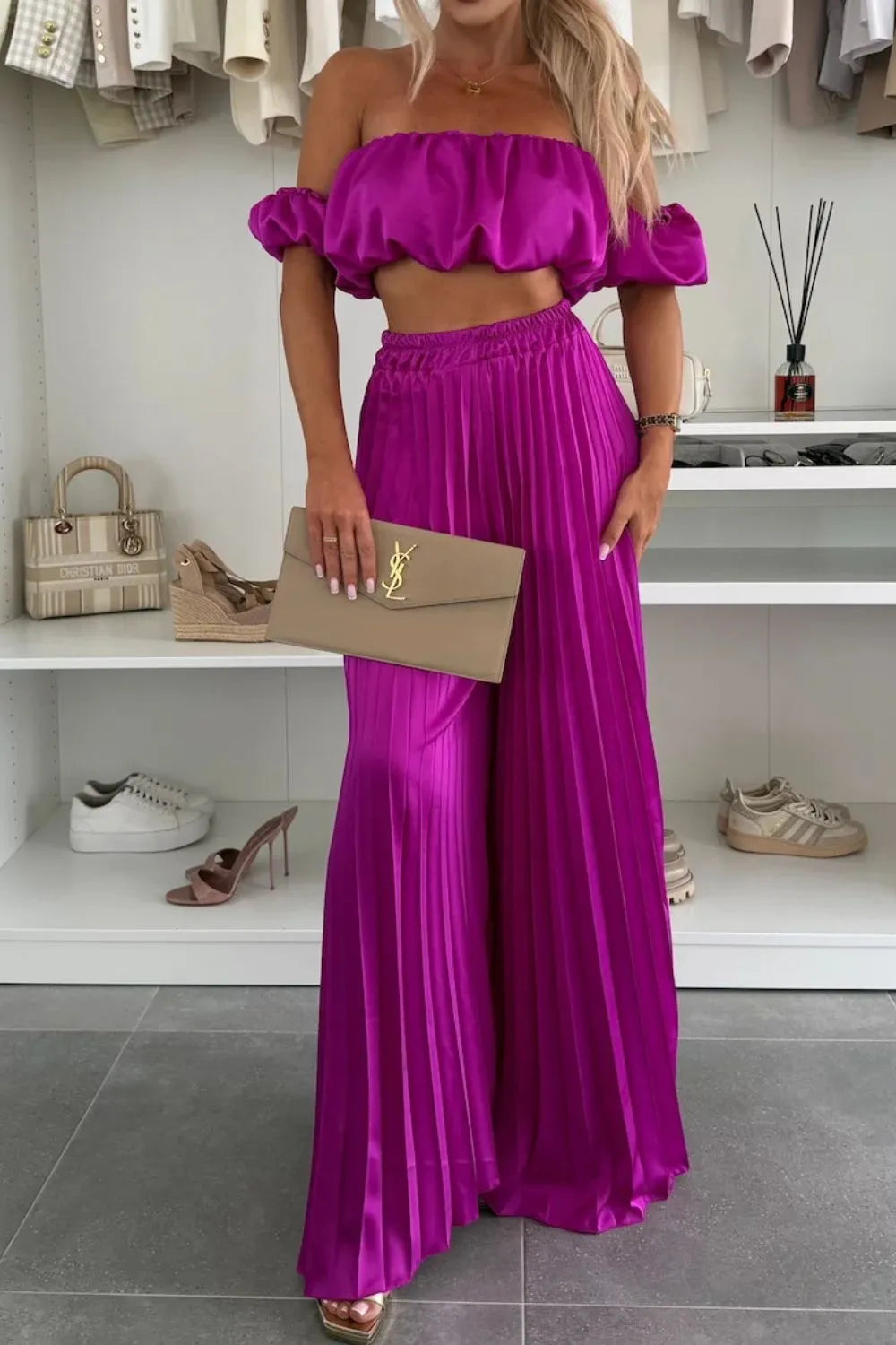 Woman wearing bright purple off-shoulder ruffled top and pleated pants set with beige clutch purse