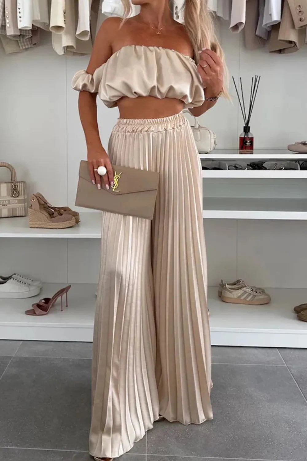 Woman wearing beige off-shoulder ruffled top and pleated wide-leg pants set holding a clutch purse in a modern closet setting