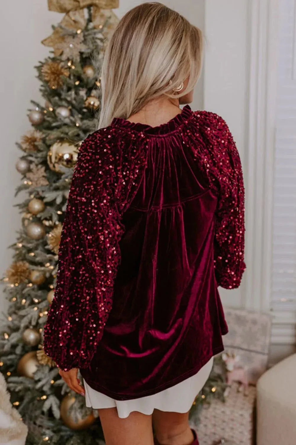 Woman wearing a burgundy sequin notched long sleeve blouse with velvet texture, posing indoors near a decorated Christmas tree.