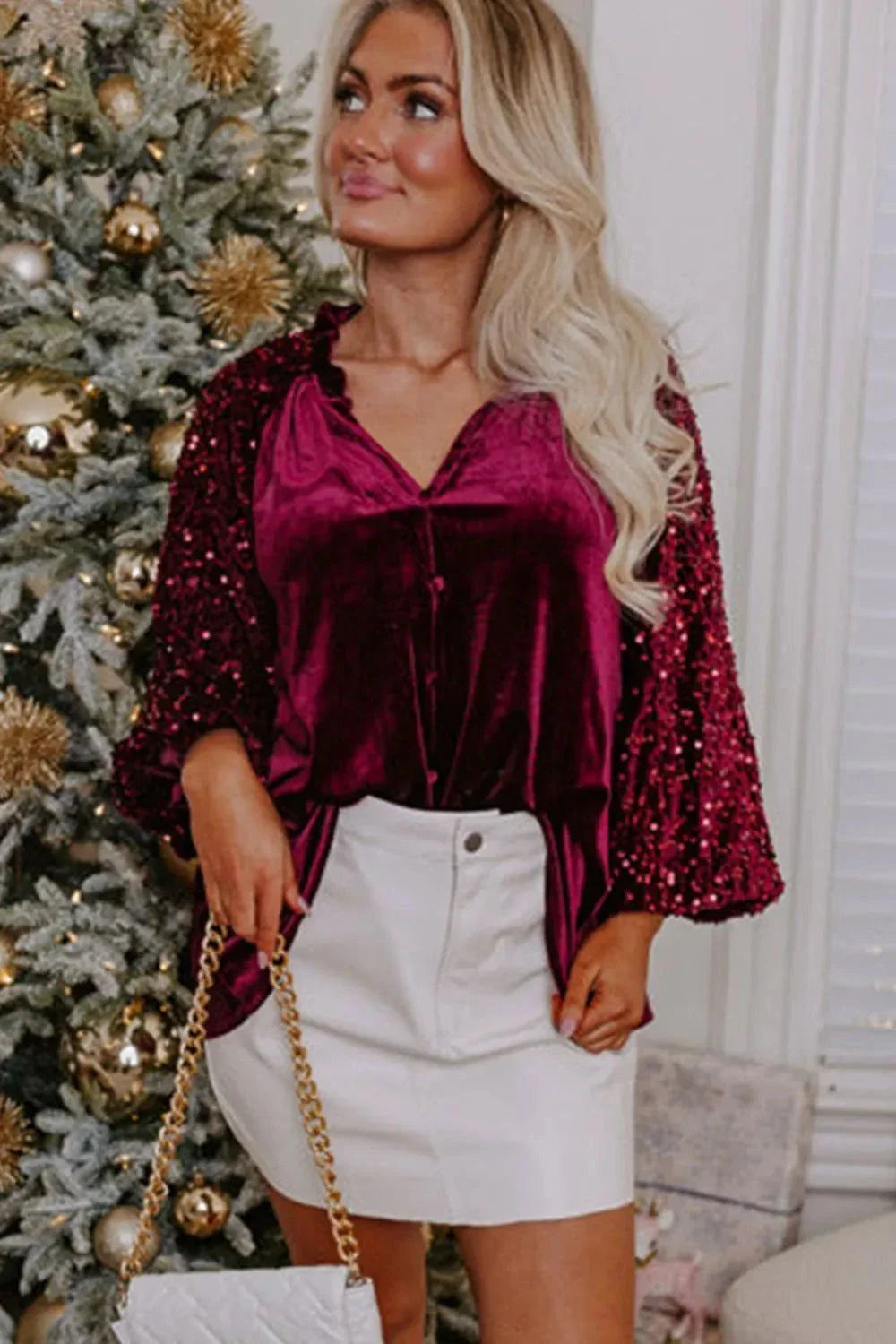 Woman wearing a burgundy sequin notched long sleeve blouse with white skirt near Christmas tree