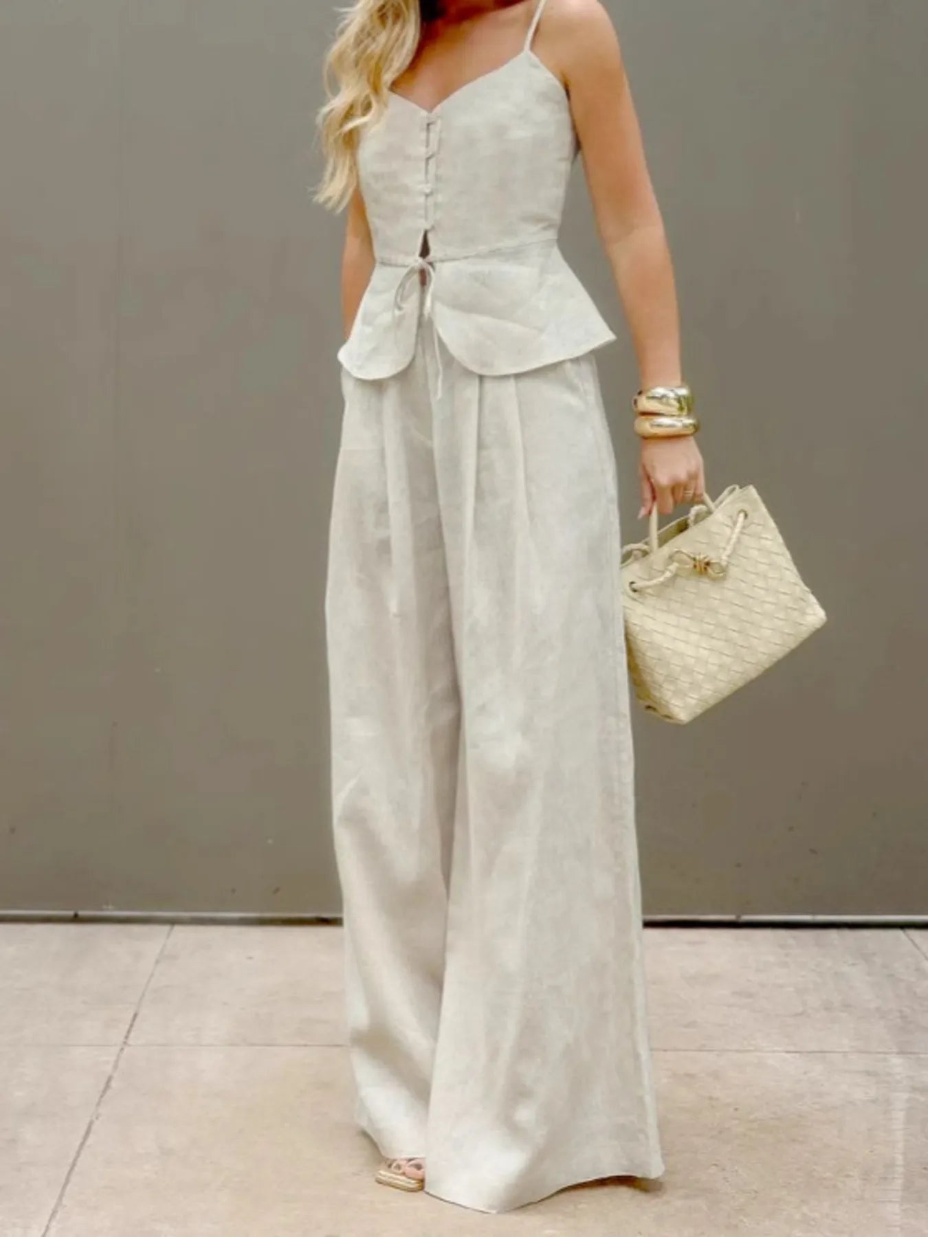 Woman wearing a beige V-neck cami and pants set with button and tie details, holding a woven handbag.